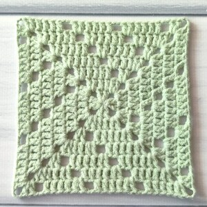 May include: A square crochet granny square in a light sage green colour. The piece features a classic openwork pattern with clusters of stitches forming a geometric design that radiates from the centre outward to the four corners of the square.