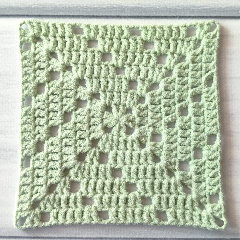 May include: A square crochet granny square in a light sage green colour. The piece features a classic openwork pattern with clusters of stitches forming a geometric design that radiates from the centre outward to the four corners of the square.