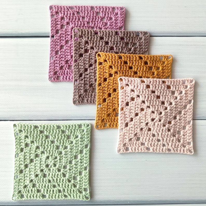 May include: Five square crochet motifs arranged in a cluster. The pieces feature a classic granny square pattern with openwork stitches. The colours include light pink, dusty rose, mustard yellow, light brown, and a soft sage green yarn.