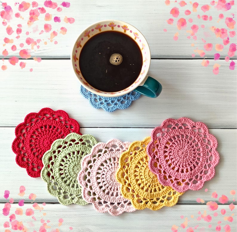May include: A cup of coffee rests on a blue crocheted coaster, with a collection of colorful, handmade coasters in red, green, pink, and yellow. The coasters are arranged on a white wooden surface, with pink floral accents.