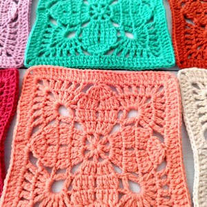 May include: Close-up of several colorful crochet squares in shades of pink, coral, green, and orange. Each square features a detailed floral pattern in the center, surrounded by a textured border. The squares are arranged closely together, showcasing the handmade craftsmanship.