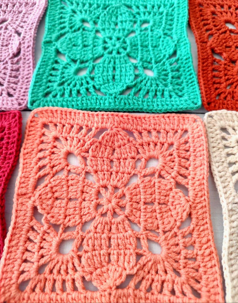 May include: Close-up of several colorful crochet squares in shades of pink, coral, green, and orange. Each square features a detailed floral pattern in the center, surrounded by a textured border. The squares are arranged closely together, showcasing the handmade craftsmanship.