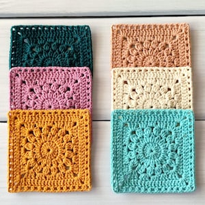 Op de afbeelding: Een verzameling van zes kleurrijke, vierkante gehaakte onderzetters. De onderzetters zijn in tinten van teal, roze, goud, perzik, crème en turquoise. Elke onderzetter heeft een bloemenontwerp in het midden. De onderzetters zijn in twee kolommen van drie gestapeld.