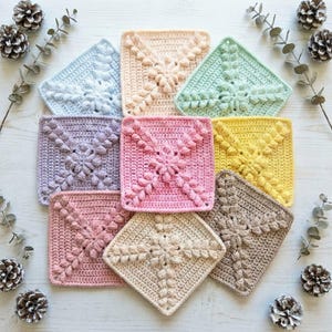 May include: Nine colourful, square crochet coasters in pastel shades. The coasters include blue, pink, yellow, and beige, each with a central floral design and textured border. The arrangement is on a white surface with pinecones and greenery, creating a cozy aesthetic.
