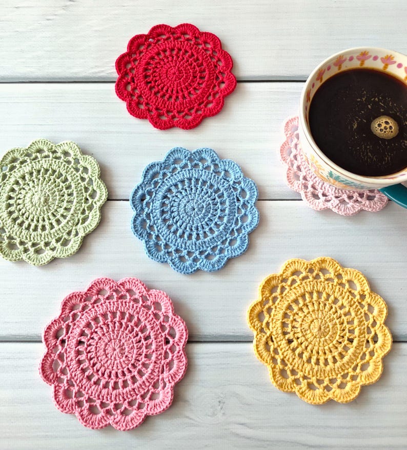 May include: A collection of colorful, handmade crochet coasters in red, green, blue, pink, and yellow. A cup of coffee rests on a pink coaster. The coasters are circular with a detailed, openwork design.