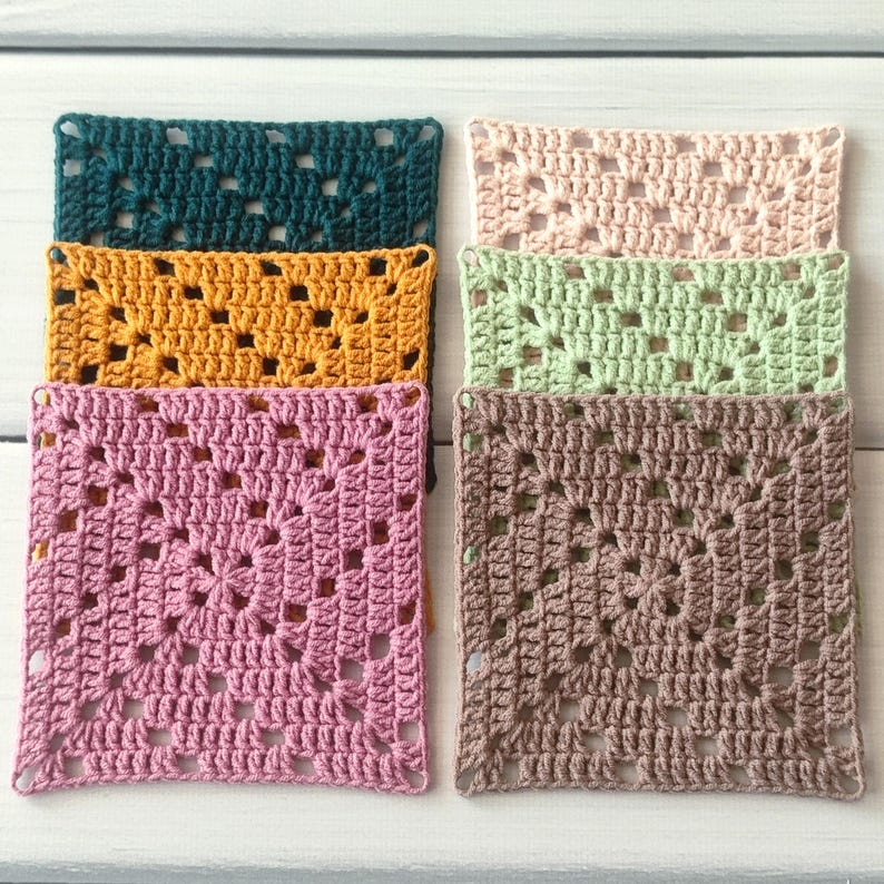 May include: A collection of six square crochet granny squares in various solid colours including dark teal, mustard yellow, dusty pink, pale peach, light sage green, and taupe. Each square features a classic openwork pattern with a central motif.
