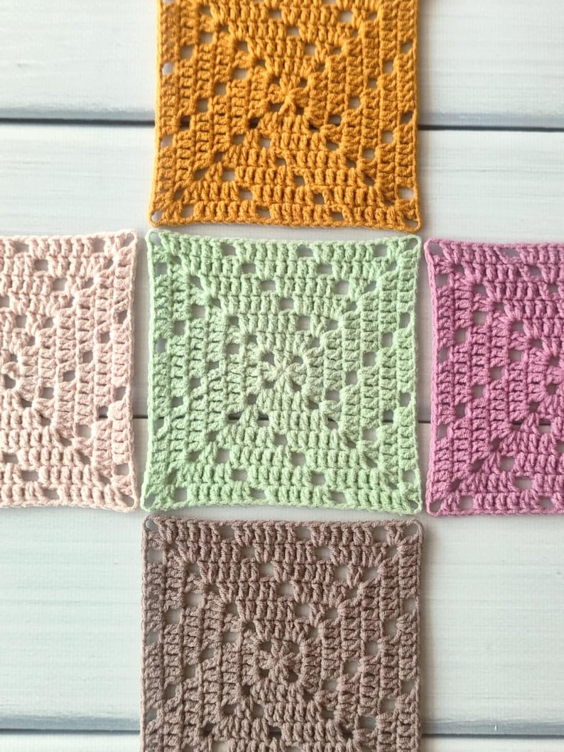 May include: Five square crochet motifs arranged in a cross pattern. The centre square is light green, surrounded by a mustard yellow square at the top, a light pink square on the left, a mauve square on the right, and a brown square at the bottom.