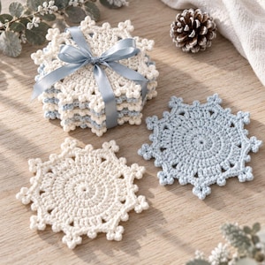 Könnte beinhalten: Ein Stapel gehäkelter Untersetzer in Schneeflockenform in Creme und Hellblau, zusammengebunden mit einem hellblauen Satinband. Zwei weitere einzelne Schneeflocken-Untersetzer, einer in Creme und einer in Hellblau, liegen separat daneben.