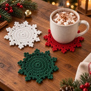 May include: Three crocheted snowflake coasters in white, red, and dark green. The red coaster supports a ceramic mug filled with hot cocoa topped with marshmallows. The coasters feature intricate, circular, star-shaped patterns with pointed edges.