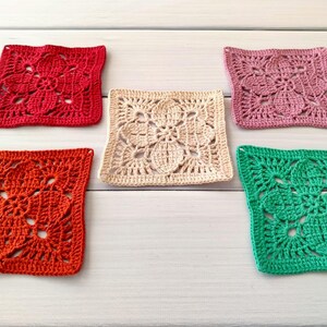May include: Five colorful, square crochet coasters. The coasters are in shades of red, orange, cream, pink, and green. Each coaster features a floral design. The coasters are arranged on a white wooden surface.