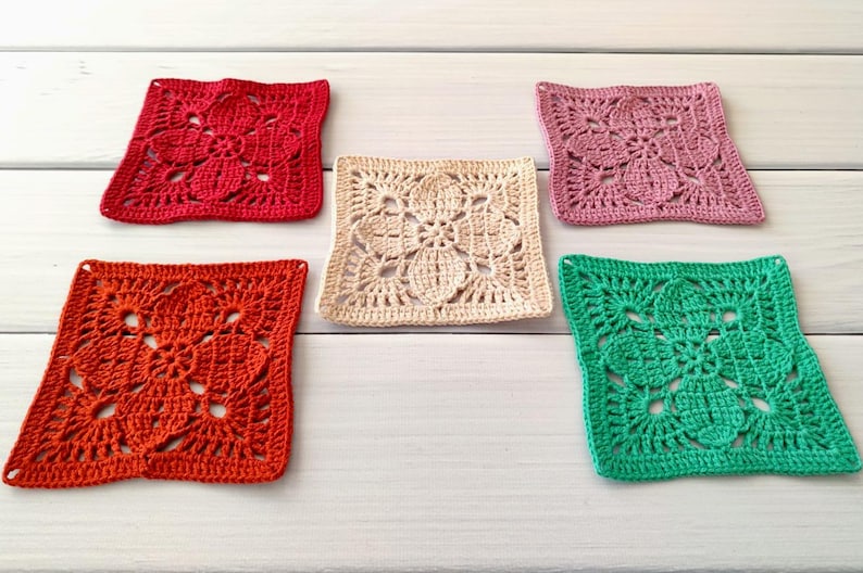 May include: Five colorful, square crochet coasters. The coasters are in shades of red, orange, cream, pink, and green. Each coaster features a floral design. The coasters are arranged on a white wooden surface.