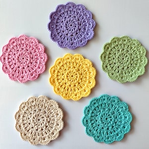 May include: A collection of six handmade crochet coasters in various colors, including pink, purple, yellow, green, beige, and teal. Each coaster features a circular design with a scalloped edge, perfect for protecting surfaces from drink condensation.