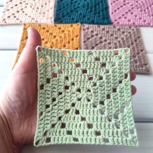 May include: A collection of six square crochet motifs in various solid colours including light green, mustard yellow, teal, light pink, dusty rose, and beige. Each piece features a central star-like pattern surrounded by openwork stitches.