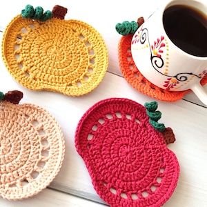 Puede incluir: Cuatro posavasos de calabaza hechos a mano con ganchillo en tonos amarillo, naranja, rosa y rojo. Cada posavasos presenta un diseño en espiral y un tallo verde y marrón. Una taza blanca con café se encuentra sobre uno de los posavasos.