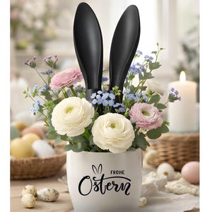 Könnte beinhalten: Ein weißer Keramiktopf mit der Aufschrift "Frohe Ostern" und einer Hasenillustration, gefüllt mit weißen, rosa und blauen Blumen und mit schwarzen Hasenohren versehen. Osterei-Blumenarrangement.