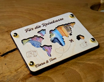 Könnte beinhalten: Eine Holz-Reisekasse mit Weltkarten-Design. Die Karte hat ausgeschnittene Kontinente, die mit bunten Währungen gefüllt sind. Die Box ist mit "Für die Reisekasse" und "Clara & Tom" beschriftet, mit goldenen Eckakzenten.