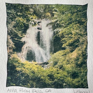 May include: A black and white photograph of Anna Ruby Falls in Georgia. The image shows a waterfall cascading down a rocky cliff, surrounded by lush green trees. The text "Anna Ruby Falls, GA" is written below the image, along with the artist's signature "J. Shaw".