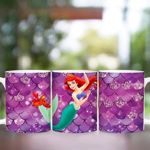 Puede incluir: Tres tazas de cer&aacute;mica blanca con un patr&oacute;n de escamas moradas e im&aacute;genes de Ariel de La Sirenita. Una taza presenta a Ariel con un cangrejo y las otras tazas muestran el patr&oacute;n de escamas con detalles brillantes.