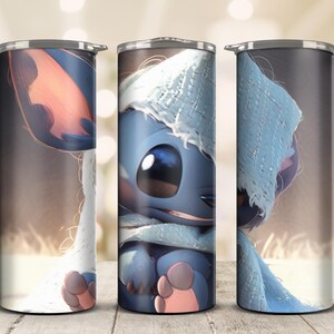 May include: A set of three stainless steel insulated tumblers. Each tumbler features a full-wrap design of a blue cartoon character wearing a light blue hat and scarf. The character has large eyes and pink ears and feet. The background is a blurred brown.
