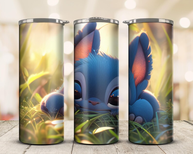 May include: A set of three stainless steel tumblers featuring a blue cartoon character in a grassy field. The character has large ears, big eyes, and a blue and pink color scheme. The background is a soft, blurred image of sunlight and greenery.