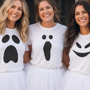 May include: Three white short-sleeved t-shirts featuring black ghost face graphics paired with white tulle skirts. Each shirt displays a unique facial expression, including a wide-eyed ghost, a surprised ghost, and a mischievous grinning ghost.