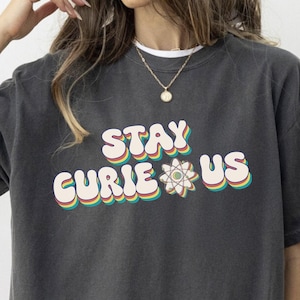 May include: Dark grey t-shirt featuring the text "STAY CURIE US" in a retro font with rainbow detailing and an atom graphic. A casual style t-shirt suitable for everyday wear.