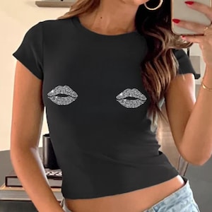 May include: Black short-sleeved crop top with two rhinestone lip designs. The top is a fitted style and is paired with light-wash denim jeans. The model is wearing sunglasses and gold hoop earrings.