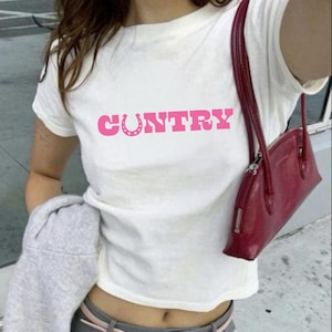Könnte beinhalten: Weißes T-Shirt mit dem Wort "COUNTRY" in rosa Blockbuchstaben und einer Hufeisen-Grafik. Eine rote Handtasche mit Schulterriemen ist zu sehen. Das Shirt wird mit grauer Hose und einem grauen Sweatshirt kombiniert.