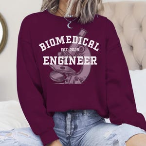 May include: Burgundy sweatshirt with a white graphic of a microscope and the text "Biomedical Engineer Est. 2025".
