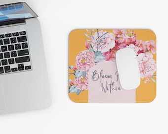 Peut inclure: Un tapis de souris rectangulaire avec un motif floral et le texte "Bloom Within". Le tapis de souris présente un fond jaune avec un arrangement de fleurs roses et blanches de style aquarelle. Une souris d'ordinateur blanche repose sur le tapis.