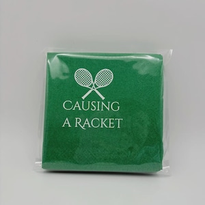 May include: A green square towel with the text "CAUSING A RACKET" and crossed tennis rackets in white. The towel is packaged in clear plastic, suitable for tennis enthusiasts.