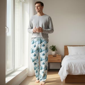 May include: A person wearing a grey long-sleeved top and patterned pyjama bottoms. The pyjama bottoms feature a white backdrop with a blue and green mountain and tree design. The person is holding a white mug.