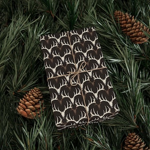 May include: A wrapped gift with a repeating pattern of white deer antlers on a dark brown background, tied with twine. The gift is surrounded by green pine needles and pine cones, creating a festive, seasonal presentation.