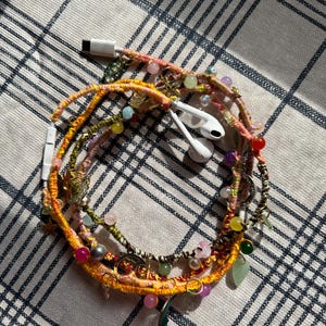 May include: A white wired earphone cable wrapped in multi-colored braided thread, featuring various small charms including stars, moons, and colorful beads in shades of pink, green, yellow, and purple, with a white USB-C connector at the end.