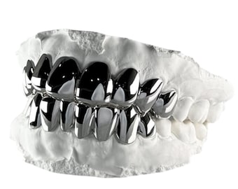 May include: A set of polished silver-coloured metal teeth grills fitted over a white plaster dental mould. The metallic covers are shaped to fit the upper and lower front teeth, featuring a smooth, reflective surface and individual tooth contours.