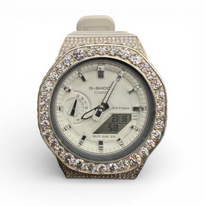 May include: A G-Shock Casio watch with a white face and a bezel encrusted with sparkling stones. The watch features a digital display showing the time, and the band is a light gray color. The watch is water resistant.