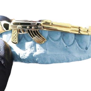 Puede incluir: Una parrilla dorada con forma de AK-47, un accesorio dental, descansa sobre un molde dental azul claro. La parrilla es muy detallada, con características visibles del arma de fuego. El fondo es blanco liso.