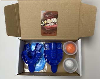 Peut inclure: Une boîte en carton ouverte contenant des gouttières d'empreintes dentaires bleues, des matériaux de moulage et un autocollant avec le texte "ICED GRILLZ" et une image de dents en or et en argent.
