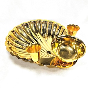 May include: A gold-colored, seashell-shaped serving dish with two small cup holders and a central bowl. The dish has a shiny, reflective surface and is set against a white background. The design is ornate and decorative.