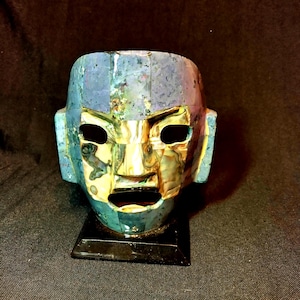 May include: A mosaic mask with a human face, featuring blue, gold, and green hues. The mask has open eye and mouth holes, and is mounted on a black stand. The mask's design suggests an ancient or cultural artifact.