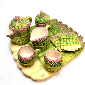 May include: A miniature tea set with a whimsical asparagus theme. The set includes a teapot, sugar bowl, creamer, and two teacups, all in shades of green and pink. The pieces sit on a leaf-shaped tray, creating a unique and charming display.