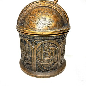 May include: A decorative, antique-style box with a globe-shaped lid and a cylindrical base. The lid features a detailed map of the world. The base is adorned with relief carvings of ships and buildings. The box is made of a bronze-colored material.