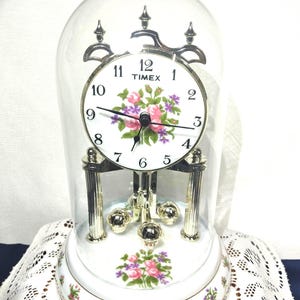 May include: A Timex clock with a white face, floral design, and silver accents. The clock is encased in a glass dome and sits on a matching floral base. The clock face displays the Timex logo and the time.
