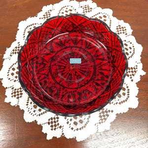 May include: A round, deep red glass bowl featuring a complex, pressed geometric pattern of diamonds and radiating lines. The bowl has a scalloped edge and a central sticker with the text "DUBLIN COLLECTION" printed in white on a teal background.