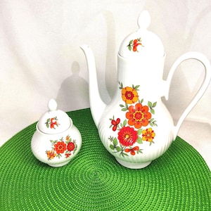 May include: White ceramic coffee pot and sugar bowl set. The coffee pot has a long spout and handle, and the sugar bowl has a lid. Both pieces are decorated with orange, red, and yellow floral patterns and green leaves.