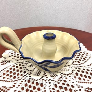 May include: Cream-colored ceramic candle holder with a blue rim and handle. The candle holder has a scalloped edge and blue leaf design. The candle holder is sitting on a white lace doily.