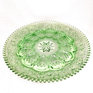May include: A round, light green glass plate featuring a central star motif surrounded by a textured diamond pattern. The border displays an intricate embossed design of flowers and scrolling vines, finished with a serrated, scalloped edge.