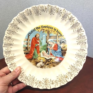 May include: Cream-colored decorative plate with a gold floral border and a central image depicting the Nativity scene. The plate features the text "Christ the Saviour is born".
