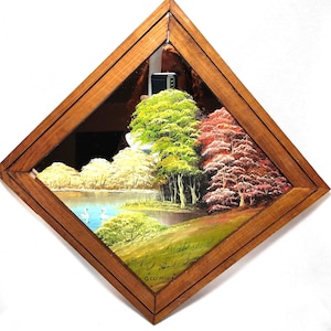 May include: A diamond-shaped mirror with a wooden frame. The mirror reflects a landscape painting of a lake, trees, and a path. The trees display green, red, and white foliage. The painting is signed in the bottom right corner.