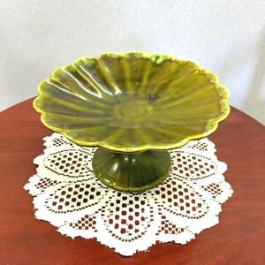 May include: A pedestal bowl featuring a scalloped edge and a glossy, olive-green glaze. The bowl has a fluted design radiating from the center, supported by a sturdy, matching circular base with vertical ridges, creating a cohesive, glazed ceramic piece.